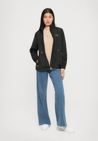 Zwarte lichtgewicht zip-up jas met verstelbare taille, elastische manchetten en gouden hardware, gecombineerd met een beige top en wijde blauwe jeans.