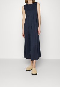 Laivaston sininen hihaton mekko, jossa on pleateja, täyspitkä malli. Yhdistetty beigen värisiin puukengiin, joissa on paksu musta pohja. Sileä kankaan rakenne.