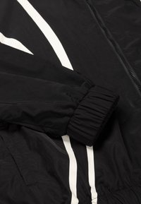 Black lightweight jacket with white vertical stripes, featuring elastic cuffs and a textured fabric. Zipper closure visible.