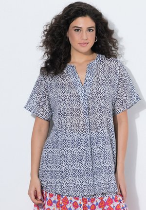 Kobieta o kręconych, ciemnych włosach, ubrana w krótki rękaw, wzorzysty biało-granatowy top oraz kolorową spódnicę z czerwonymi i pomarańczowymi abstrakcyjnymi kształtami.