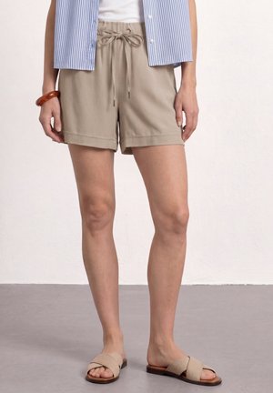 Person iført beige snørede shorts, en blå- og hvidstribet skjorte, et rødt armbånd og beige slip-on sandaler, stående på en grå gulv.
