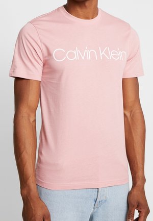 Mężczyzna w jasnoróżowej koszulce z krótkim rękawem z logo "Calvin Klein" w białym kolorze na wysokości klatki piersiowej oraz w jasnoniebieskich dżinsach.