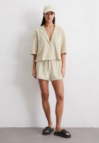 Camicia di lino in beige chiaro con maniche corte, colletto e tasca; pantaloni corti abbinati. Ciabatte nere texturizzate completano l'outfit.