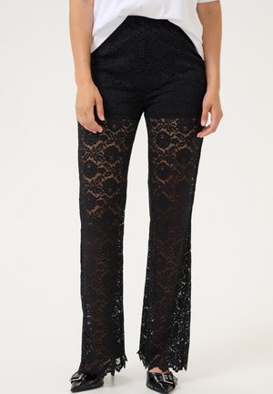 Schwarze Spitzenhose mit floralen Mustern, lockerer Passform und ausgefransten Säumen. Getragen mit einem weißen Oberteil und schwarzen Schuhen. Leichte, atmungsaktive Textur.