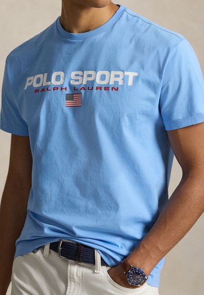 Ανοιχτό μπλε βαμβακερό μπλουζάκι με λευκό και κόκκινο κείμενο "POLO SPORT RALPH LAUREN" και μικρό γραφικό της αμερικανικής σημαίας. Κοντά μανίκια, στρογγυλή λαιμόκοψη.