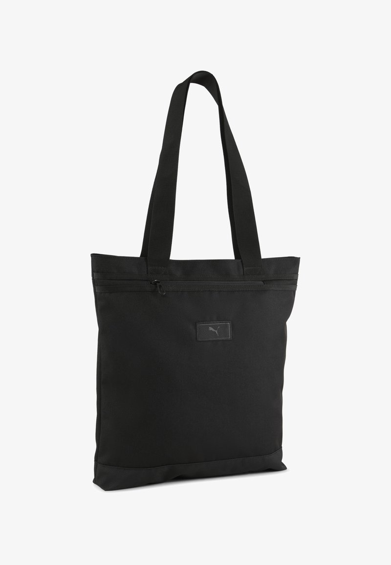 Geantă tote neagră fabricată dintr-un material durabil. Dispune de două mânere pentru umăr, un capac cu fermoar și o mică etichetă cu logo în partea din față.