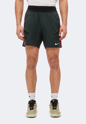 Homme portant un short de sport noir avec des bandes rouges, des chaussettes blanches, des baskets beiges et noires, debout devant un fond uni.