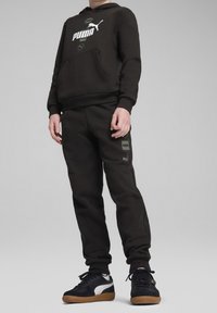 Černá mikina s kapucí a bílým a zeleným logem Puma, přední kapsa; odpovídající černé tepláky s bočními akcenty. Nosené s černými a bílými teniskami.