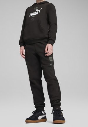 Černá mikina s kapucí a bílým a zeleným logem Puma, přední kapsa; odpovídající černé tepláky s bočními akcenty. Nosené s černými a bílými teniskami.