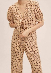 Mono floral de tela marrón, con un lazo en la parte frontal, mangas cortas abullonadas y un patrón repetido de margaritas amarillas y blancas.