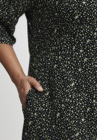 Rochie neagră cu un model stropit în verde și alb, având o cusătură la talie și un buzunar lateral, cu o siluetă liberă și fluentă și mâneci scurte.