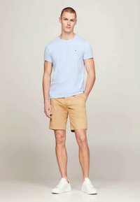 Tânăr stând cu mâinile în buzunare, purtând un tricou albastru deschis, pantaloni scurți bej și adidași albi pe un fundal simplu.