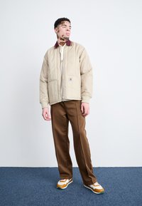 Beige gesteppte Jacke mit burgunderfarbenem Kragen, kombiniert mit braunen Hosen. Das Modell trägt weiße und orange Sneakers. Einfaches Design, minimale Muster.