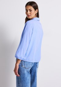 Blusa azzurro chiaro a maniche lunghe con colletto e polsini arricciati, abbinata a jeans in denim blu sbiadito a vestibilità ampia. Texture semplice e liscia.