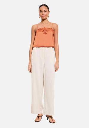 Kvinde iført ermeløs rustorange broderet top, vidde-ben beige bukser, guldringe og sorte sandaletter med hæl, stående mod hvid baggrund.