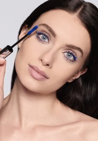 Niebieski tusz do rzęs jest nakładany za pomocą szczoteczki na rzęsy. Modelka ma proste, ciemne włosy, jasną cerę i subtelny kolor ust. Tło jest jasnoszare.