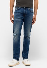 Blå denimjeans med rak ben, med ett rivet knädetalj och ljus blekning. Bärs med vita sneakers.