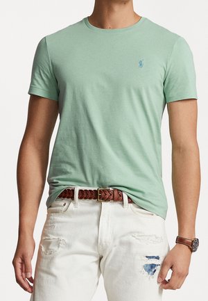 Lichtgroen katoenen t-shirt met korte mouwen, ronde halslijn en een klein blauw logo op de borst, gecombineerd met versleten witte jeans en een bruine gevlochten riem.