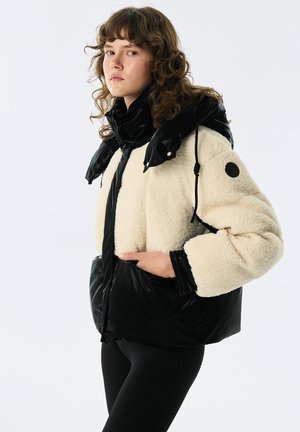 Veste en fausse fourrure noire et crème, dotée d'un col haut, d'une fermeture éclair et de grandes poches. Les accents incluent un logo rond noir sur la manche.