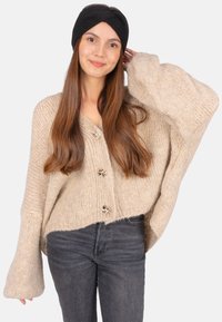 Cardigan en tricot beige avec de grandes manches et quatre boutons décoratifs. Assorti à un jean gris foncé et un bandeau noir. Texture lisse.