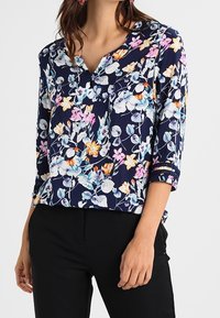 Blusa floreale con sfondo blu navy e fiori vivaci rosa, arancio e blu, scollatura a V e maniche a tre quarti.