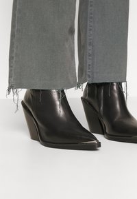 Zwarte leren wedge enkellaarsjes met een puntige neus en metallic accenten, gecombineerd met grijze cropped jeans met rafelige zomen.