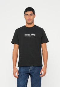 Jaunas vyras dėvi juodą Calvin Klein marškinėlius ir mėlynas džinsines kelnes, stovintis priešais vientisą baltą foną.