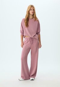 Giovane donna in piedi che indossa una felpa rosa polverosa e pantaloni larghi con sneakers bianche su uno sfondo chiaro semplice.