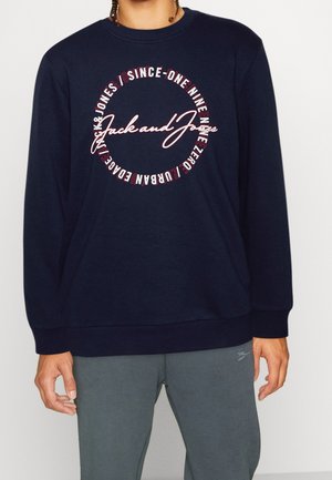 Osoba ubrana w granatową bluzę z napisem „Jack and Jones” w okrągłym wzorze, połączoną z szarymi spodniami dresowymi.