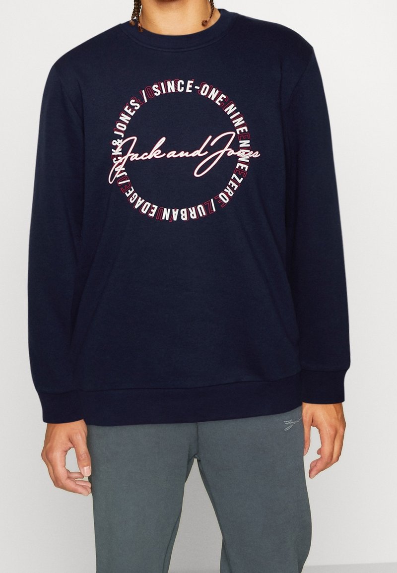 Personne portant un sweat-shirt bleu marine avec le texte « Jack and Jones » à l'intérieur d'un design circulaire, associé à un pantalon de survêtement gris.
