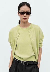 Cardigan verde chiaro sopra un maglione a girocollo abbinato, entrambi realizzati in morbido tessuto lavorato a maglia. Cintura nera con una fibbia metallica. Occhiali da sole scuri.