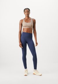 Sports-BH i lys brun med logo, kombinert med marineblå høytaljede leggings. Modellen har på seg beige treningssko med teksturerte såler.