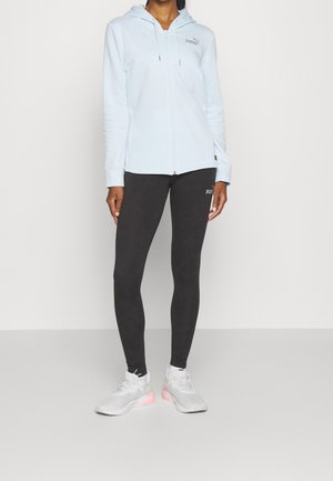 Osoba ubrana w jasnoniebieską bluzę Puma zapinaną na zamek, czarne legginsy Puma i białe sneakersy z różowymi akcentami, stojąca na tle jednolitego tła.