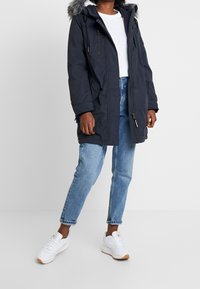 Navy parka with a fur-lined hood, featuring a front zipper, side pockets, and a relaxed fit. Worn over a white shirt and blue jeans.