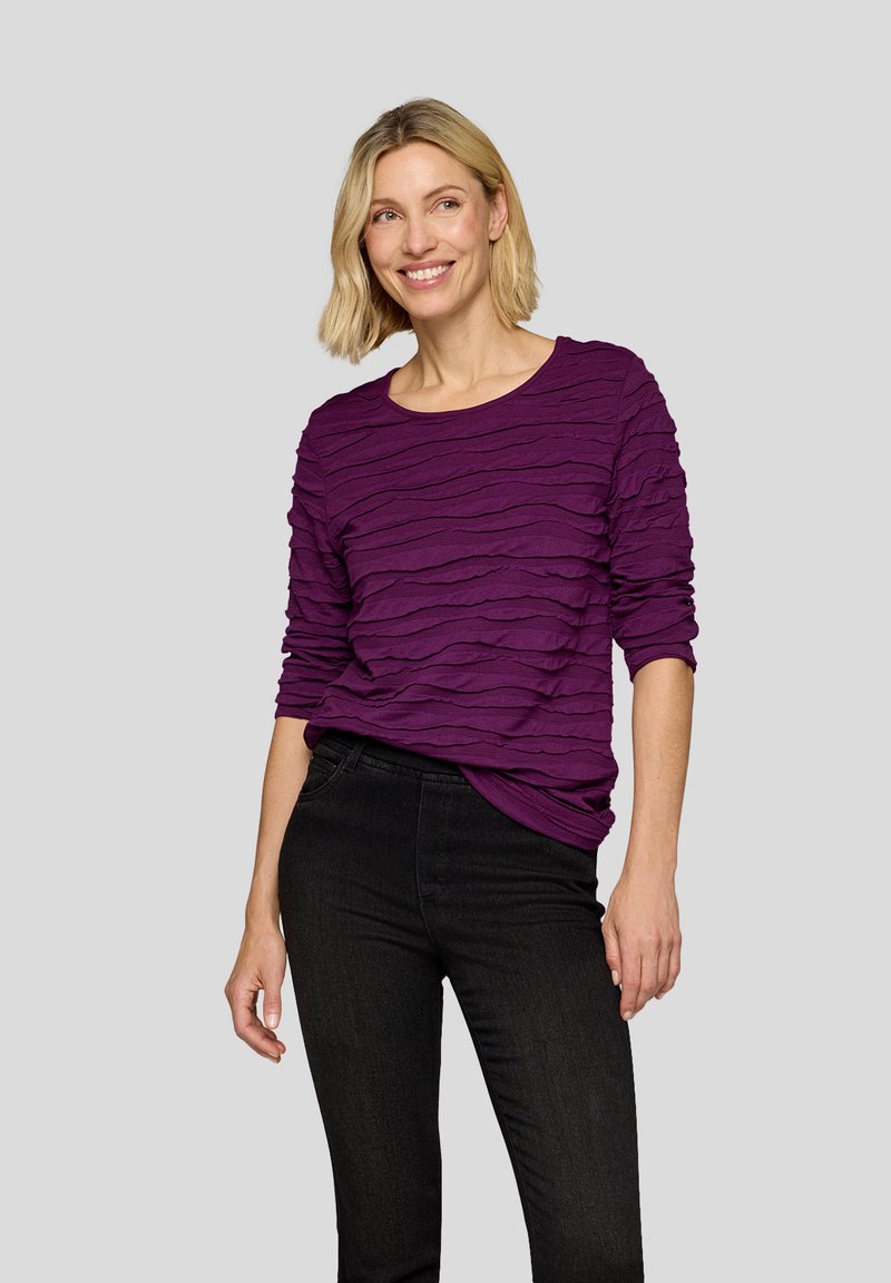 Lila Langarmoberteil mit einer kräuselförmigen Textur, rundem Ausschnitt und lockerer Passform, kombiniert mit schwarzen, eng anliegenden Jeans.