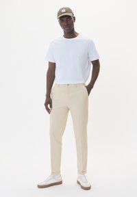 Herrklädsel bestående av en enkel vit t-shirt, beige byxor och vita sneakers med beige detaljer. Kappen är olivgrön med en logotypapplikation.