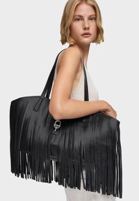 Mujer con cabello rubio corto, vestida con una blusa sin mangas, lleva un gran bolso tote de cuero negro con flecos sobre su hombro, con un fondo liso.