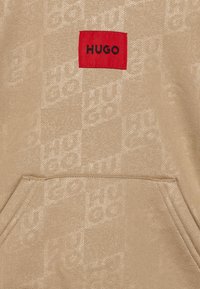 Felpa beige con una texture morbida, caratterizzata da un'etichetta rettangolare rossa con "HUGO" in nero. Include una tasca frontale e un motivo logo sottile.