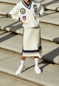 Sweat-shirt blanc avec écussons, col en V et bordure marine ; jupe midi côtelée blanche avec accents marines ; baskets blanches et chaussettes.