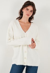 Cardigan alb, pufos, cu decolteu adânc în formă de V, mâneci lungi și șase butoane acoperite cu țesătură pe față. Textură moale și croială lejeră.