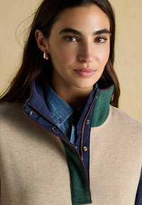 Béžový fleeceový pulóver s vysokým golierom a zelenými akcentmi, s tmavomodrou vnútornou vrstvou. Zapínanie na zips so kontrastným prešívaním.