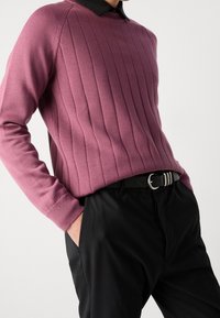 Homme portant un pull côtelé rose par-dessus une chemise noire à col, un pantalon noir avec ceinture noire, debout les mains dans les poches.