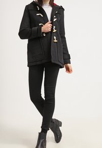 Cappotto duffle nero con cappuccio, chiusure a toggles e tasche frontali. Indossato sopra un top grigio e jeans skinny neri. Stivali neri alla caviglia con suole scanalate.