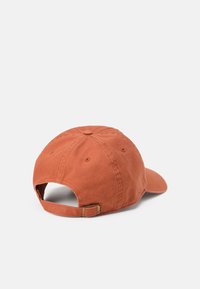 Cappello da baseball in cotone di colore ruggine con visiera curva, cinturino regolabile sul retro e un bottone sulla parte superiore. Presenta dettagli cuciti.