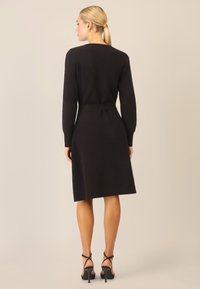 Schwarzes Strickkleid mit langen Ärmeln, tailliertem Schnitt und ausgestelltem Rock. Kombiniert mit schwarzen Riemchen-Heels. Glatte Textur, minimalistisches Design.