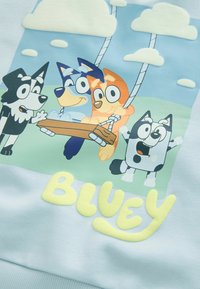 Vier tekenfilmhonden met grote ogen spelen buiten; een hond schommelt op een houten schommel die hangt aan wolken die op bomen lijken. Het woord "Bluey" staat eronder.