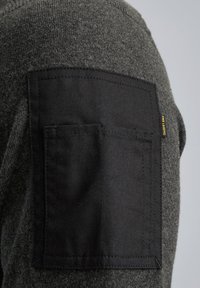 Dunkelgrauer Strickpullover mit strukturierter Oberfläche, der auf der linken Schulter ein großes schwarzes Stoffpatch mit einem kleinen gelben Etikett hat.