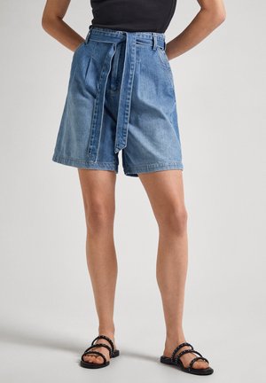 Kvinde iført lyseblå denimshorts med høj talje og bindebælte, sort ærmeløs top og sorte flade sandaler med remme.