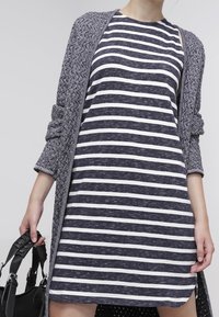 Niebiesko-biała sukienka w paski z okrągłym dekoltem, zestawiona z teksturowanym szarym kardiganem. Modelka trzyma czarną torebkę.