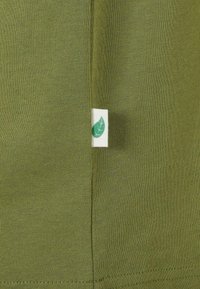 Cămașă din bumbac de culoare verde măslinie, cu o textură ribbed și o etichetă mică albă cu un simbol de frunză verde pe șold.